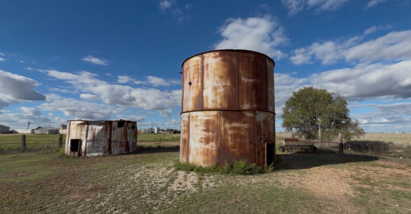 Rusty abandoned metal water tank and old shed on rural farmland under a blue sky with white clouds.