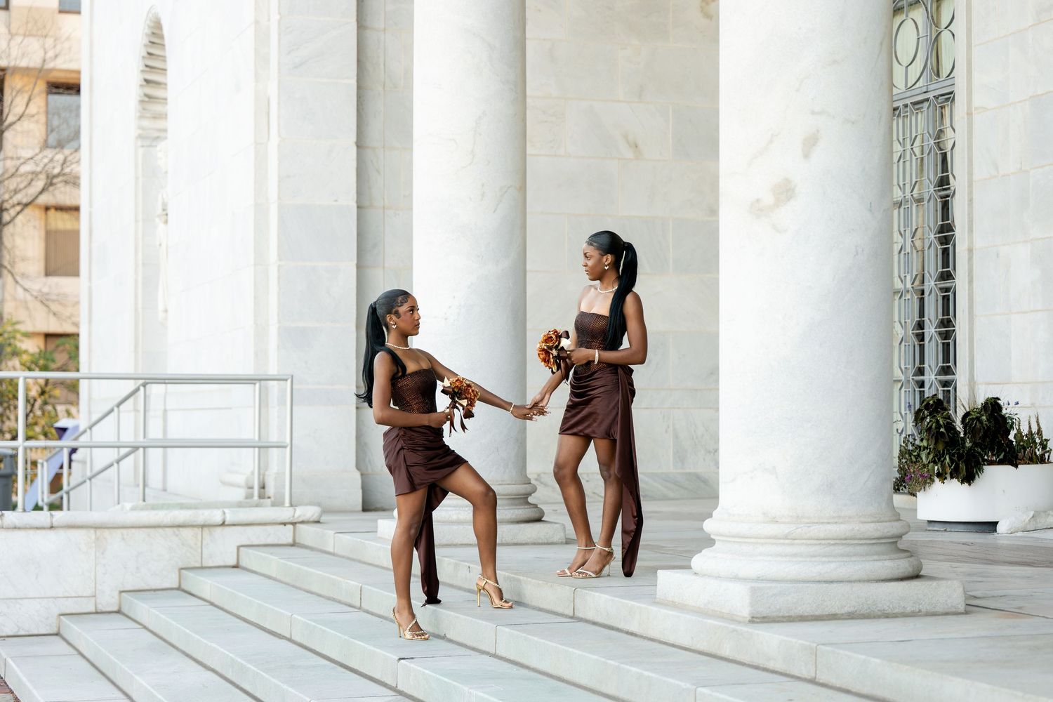 Two women in brown dresses hold bouquets while posing on stone steps between tall white columns of a classical building.