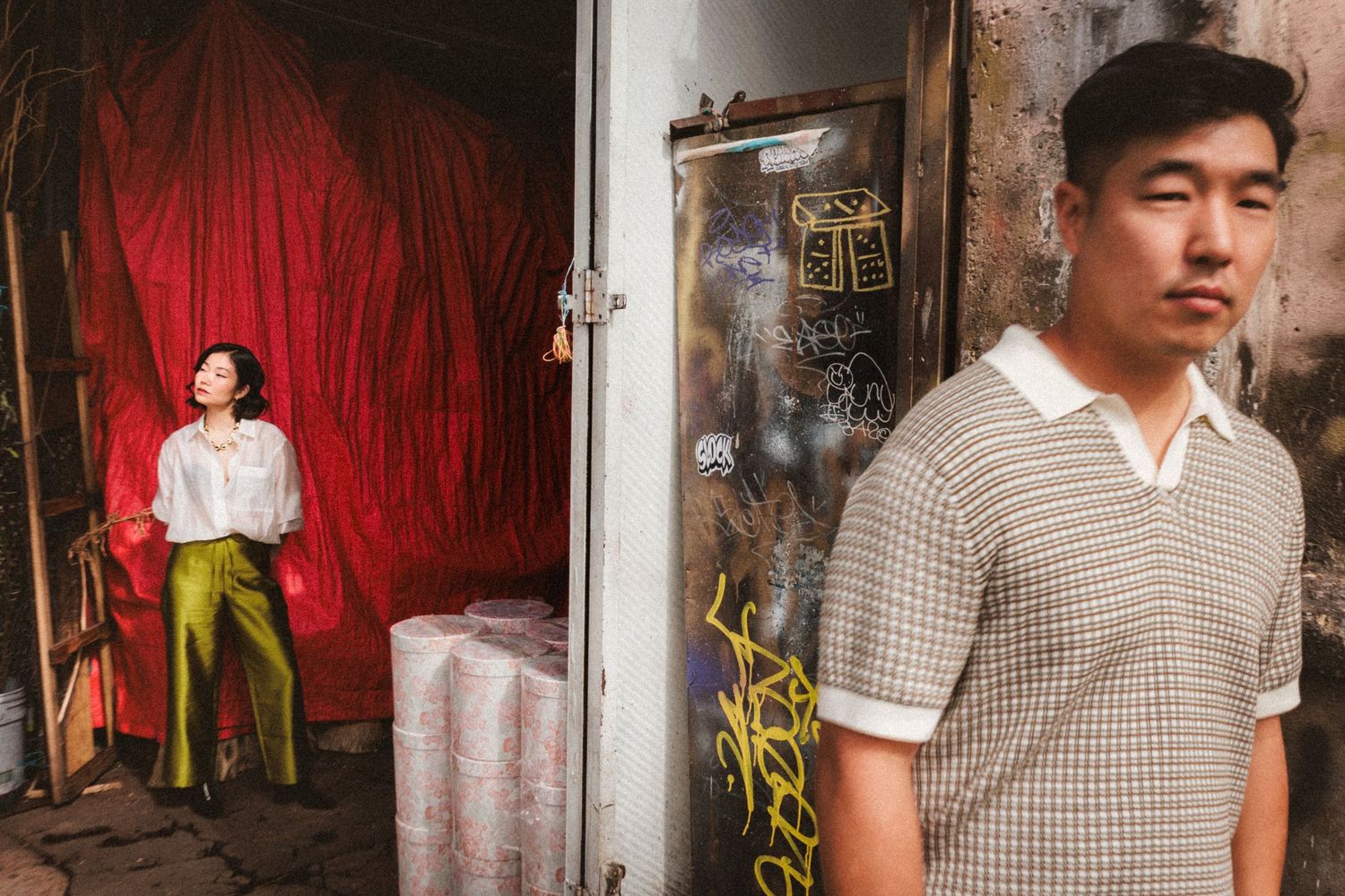 A man in a plaid shirt stands near a graffiti wall while a woman in green pants poses by a red curtain.