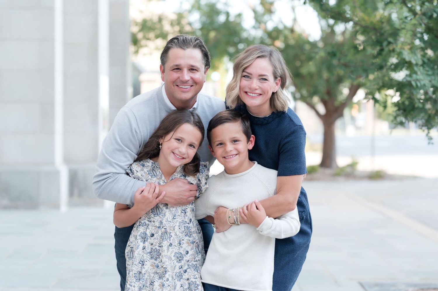 Happy family of four smiling outdoors, parents hugging two young children in a bright urban setting.