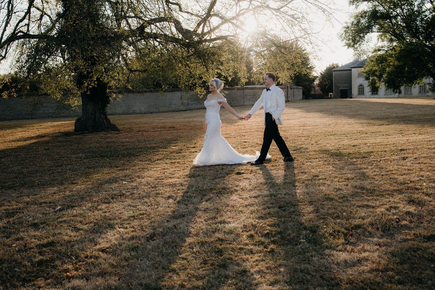 Bride and groom holding hands walking through sunlit park grounds in elegant wedding attire.