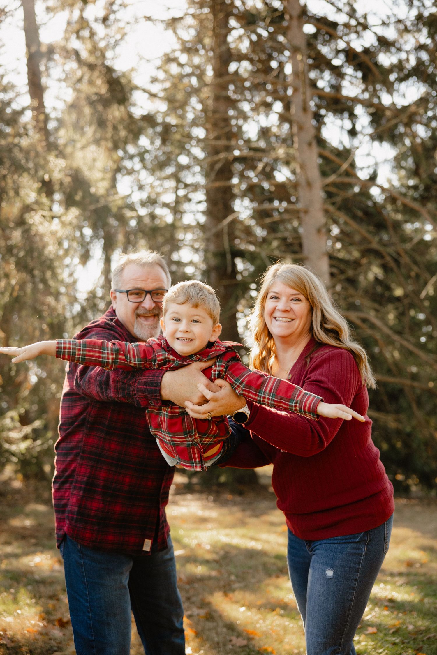 A joyful family of three in red plaid outfits poses outdoors among tall pine trees on a sunny autumn day.