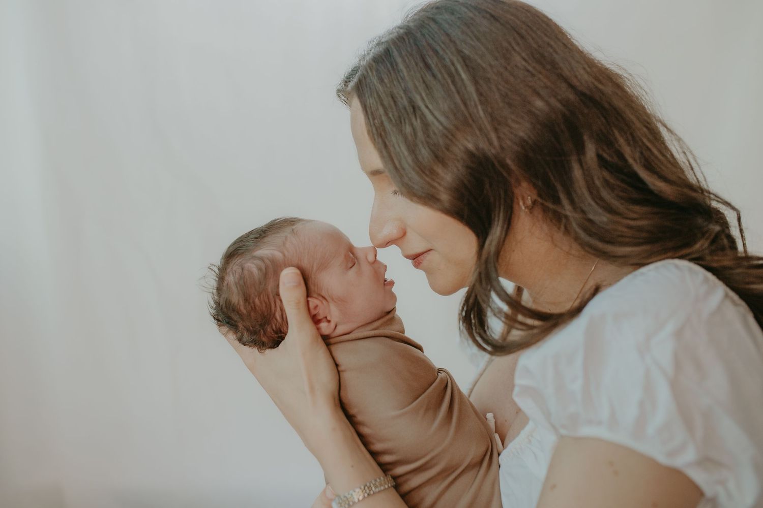A mother tenderly holds her swaddled newborn baby close, touching noses in an intimate bonding moment.
