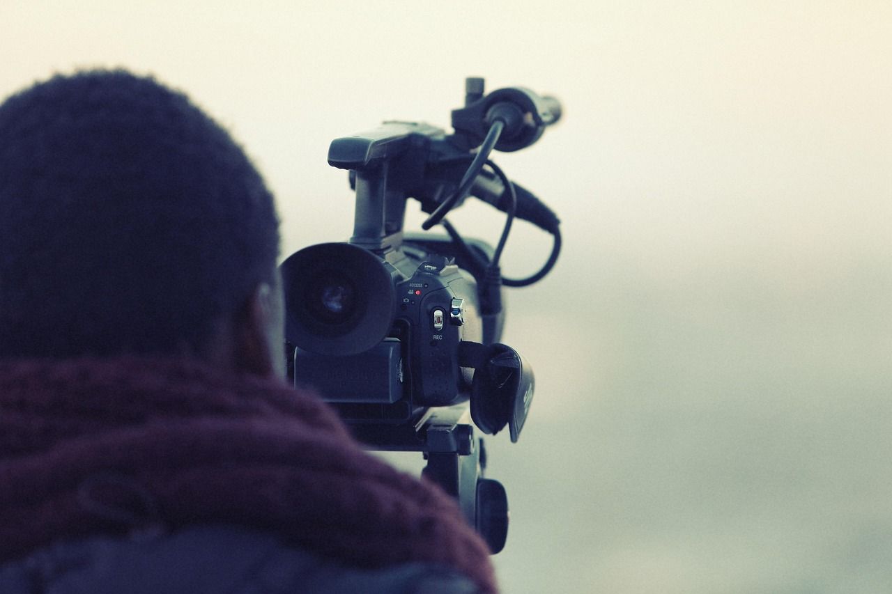 Professional camera operator filming with a broadcast video camera and wearing a burgundy hoodie in outdoor setting.