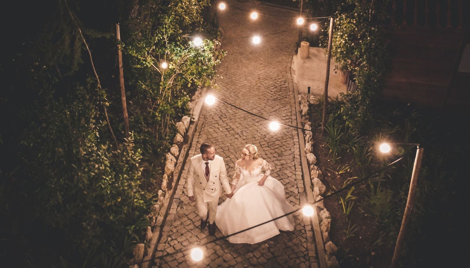 Aerial view of bride and groom walking down a cobblestone path lit by string lights at night.