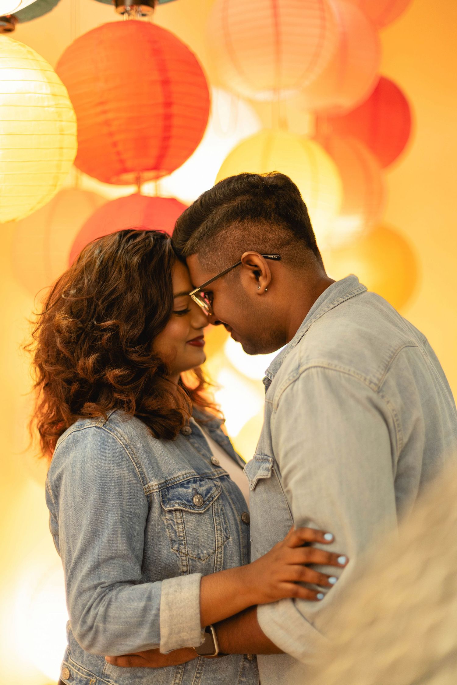 A romantic couple in denim jackets embracing tenderly with colorful paper lanterns glowing behind them.
