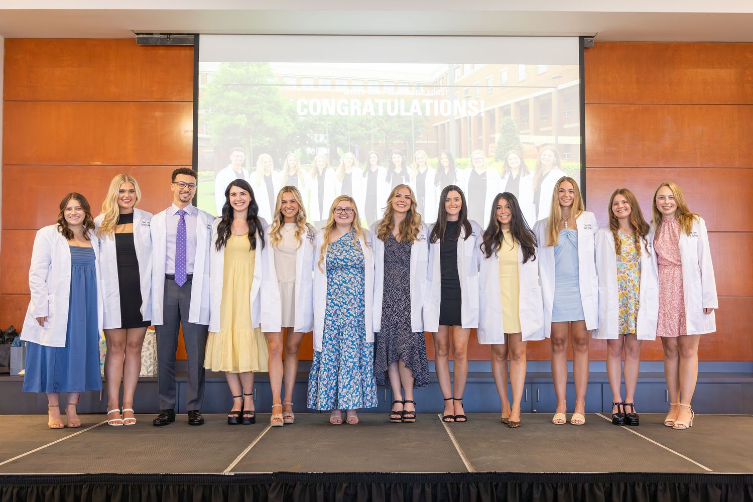 A row of students stand together wearing white medical coats during a professional ceremony event.