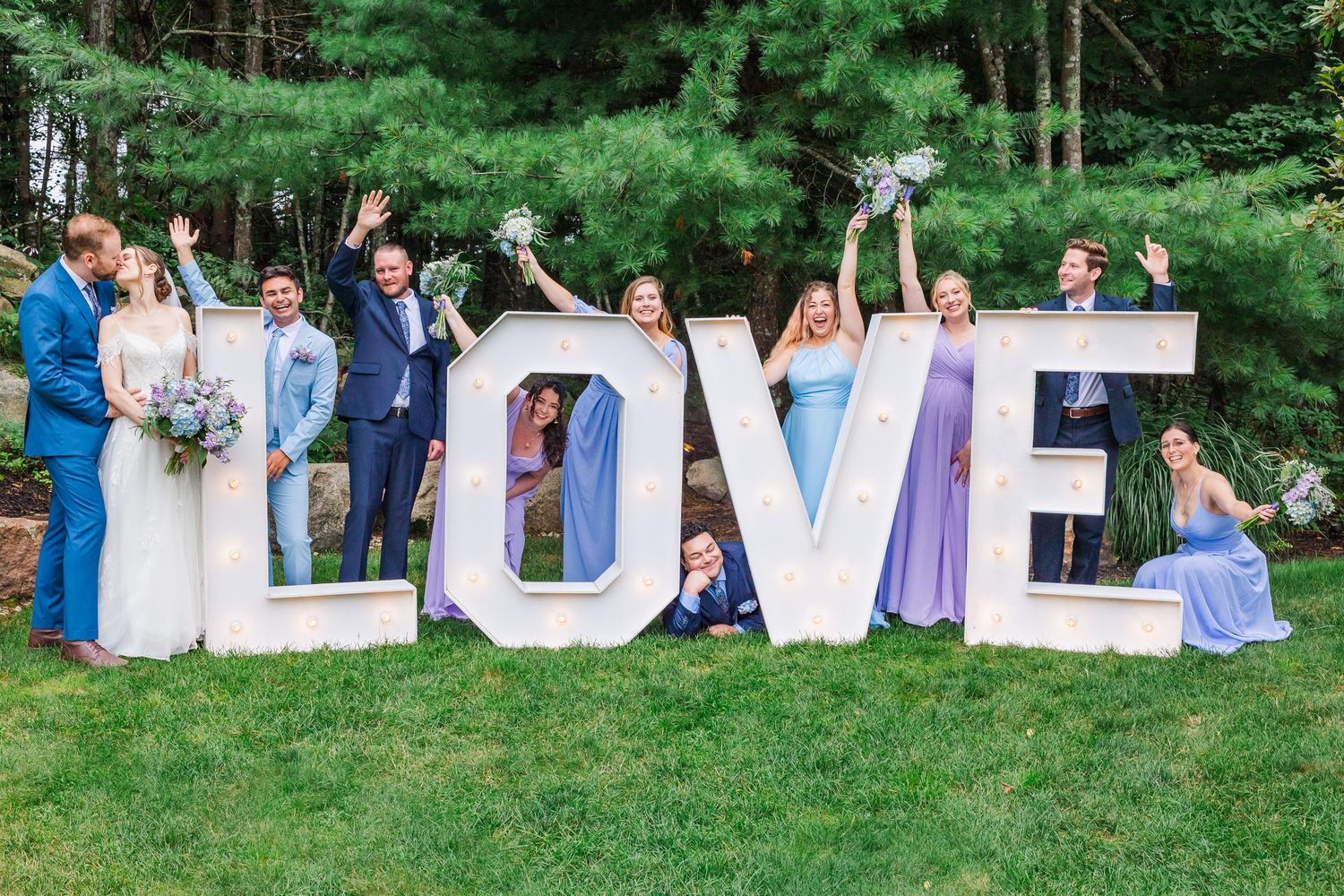 Wedding party members pose with giant white LOVE letters outdoors among green pine trees at outdoor celebration.