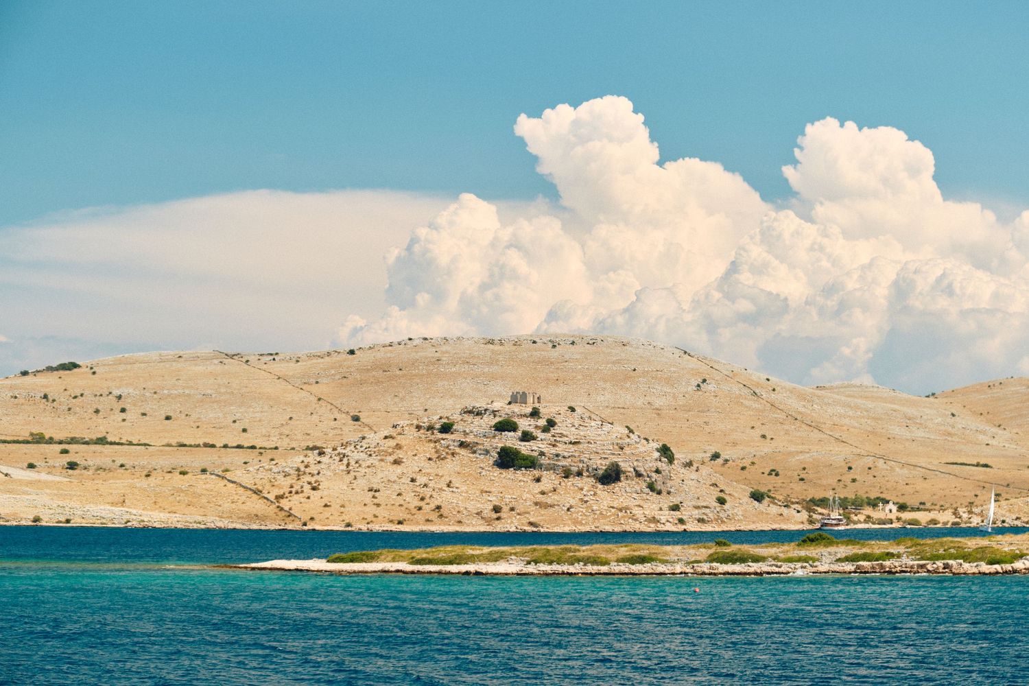 Barren coastal hills rise above turquoise waters with dramatic white clouds in background.
