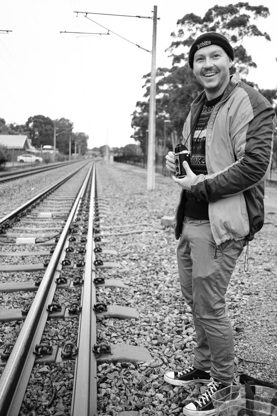 Photographer standing along train tracks with camera and casual attire in black and white composition.