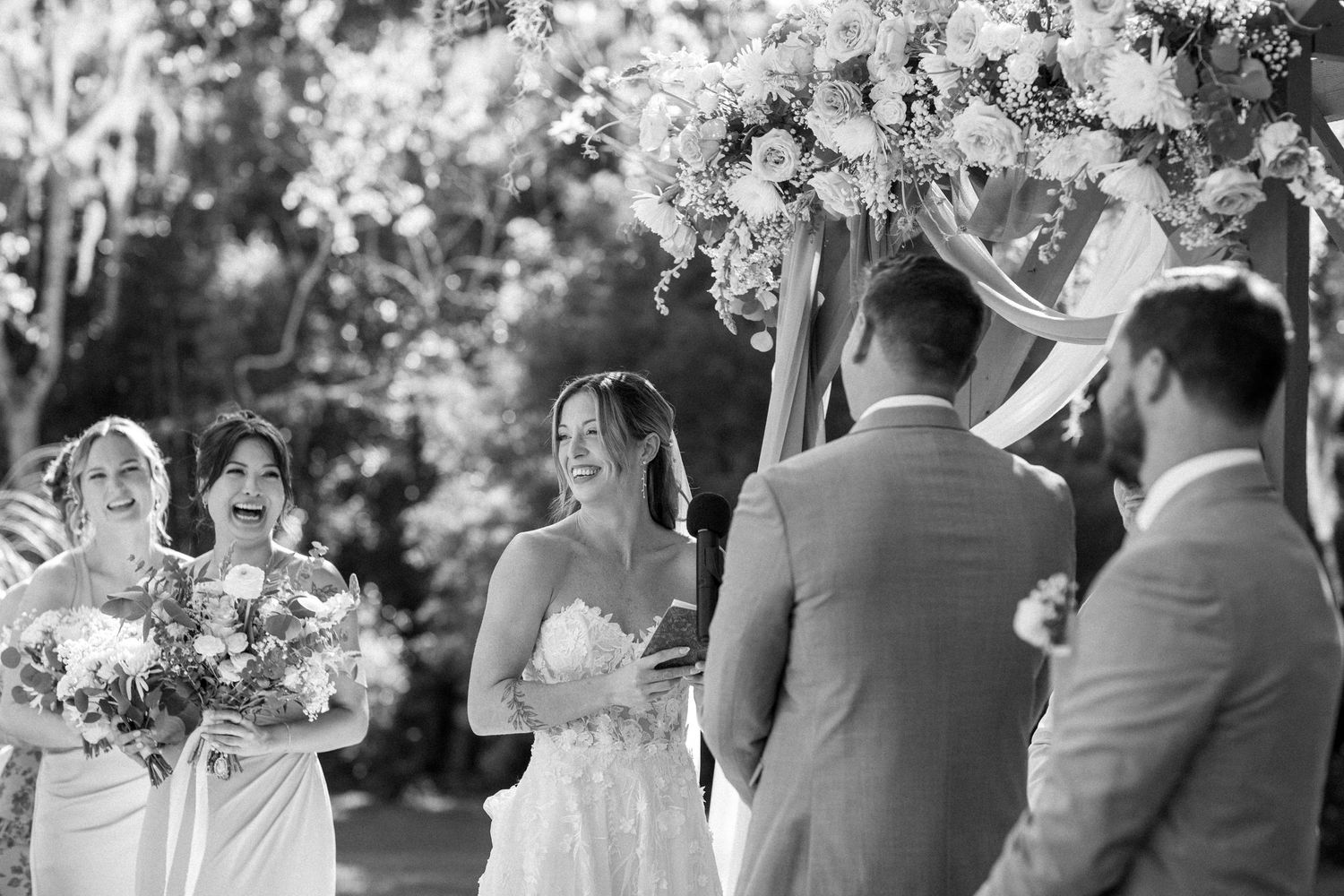 Black and white photo of an outdoor wedding ceremony under a floral arch with guests gathered around.