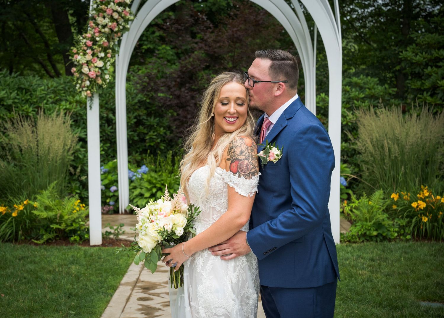A romantic wedding couple embraces under a white arch in a garden setting with a flowing veil creating a beautiful frame.