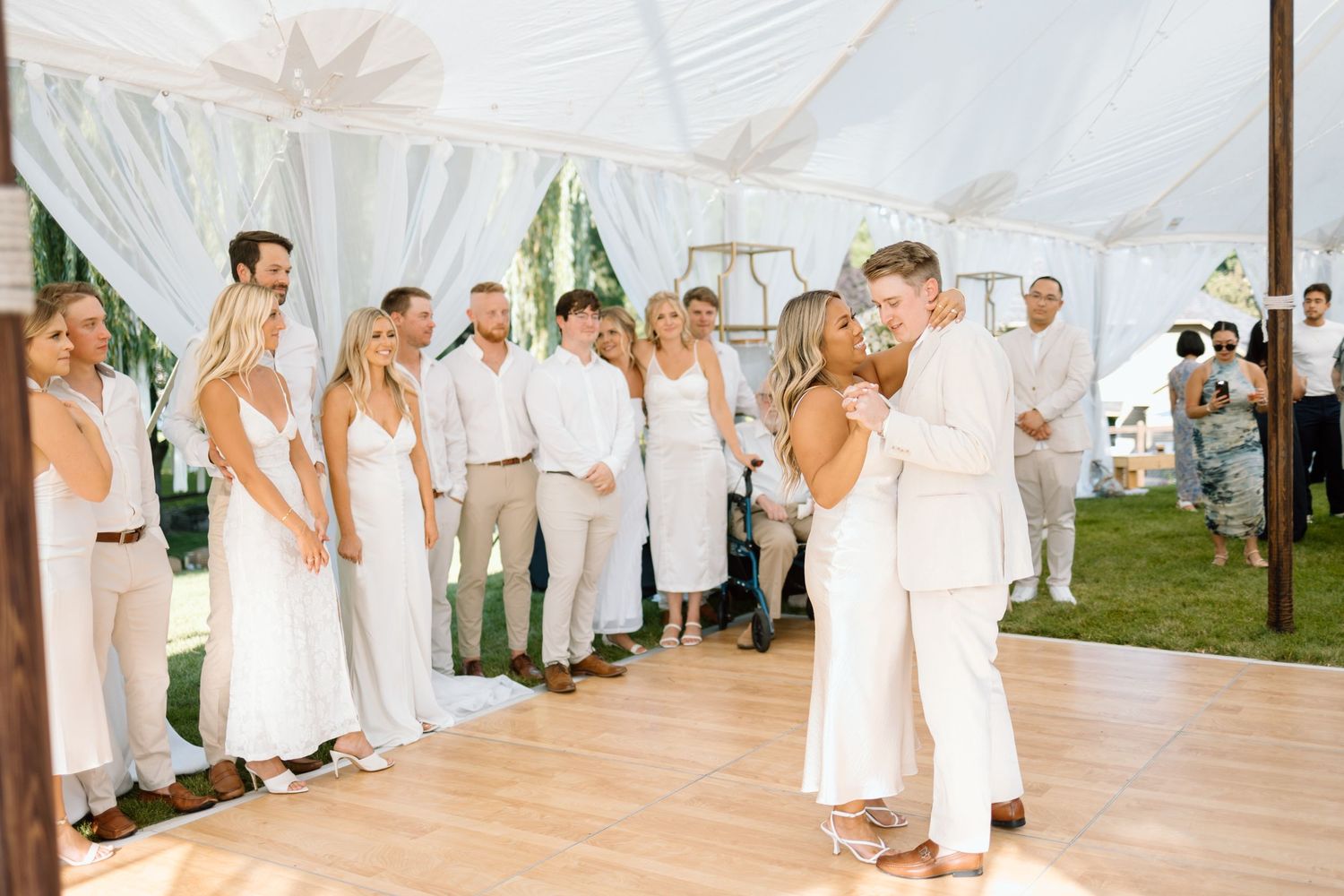 People in white attire gather at an elegant outdoor tent wedding reception as a couple shares a dance on a wooden floor.