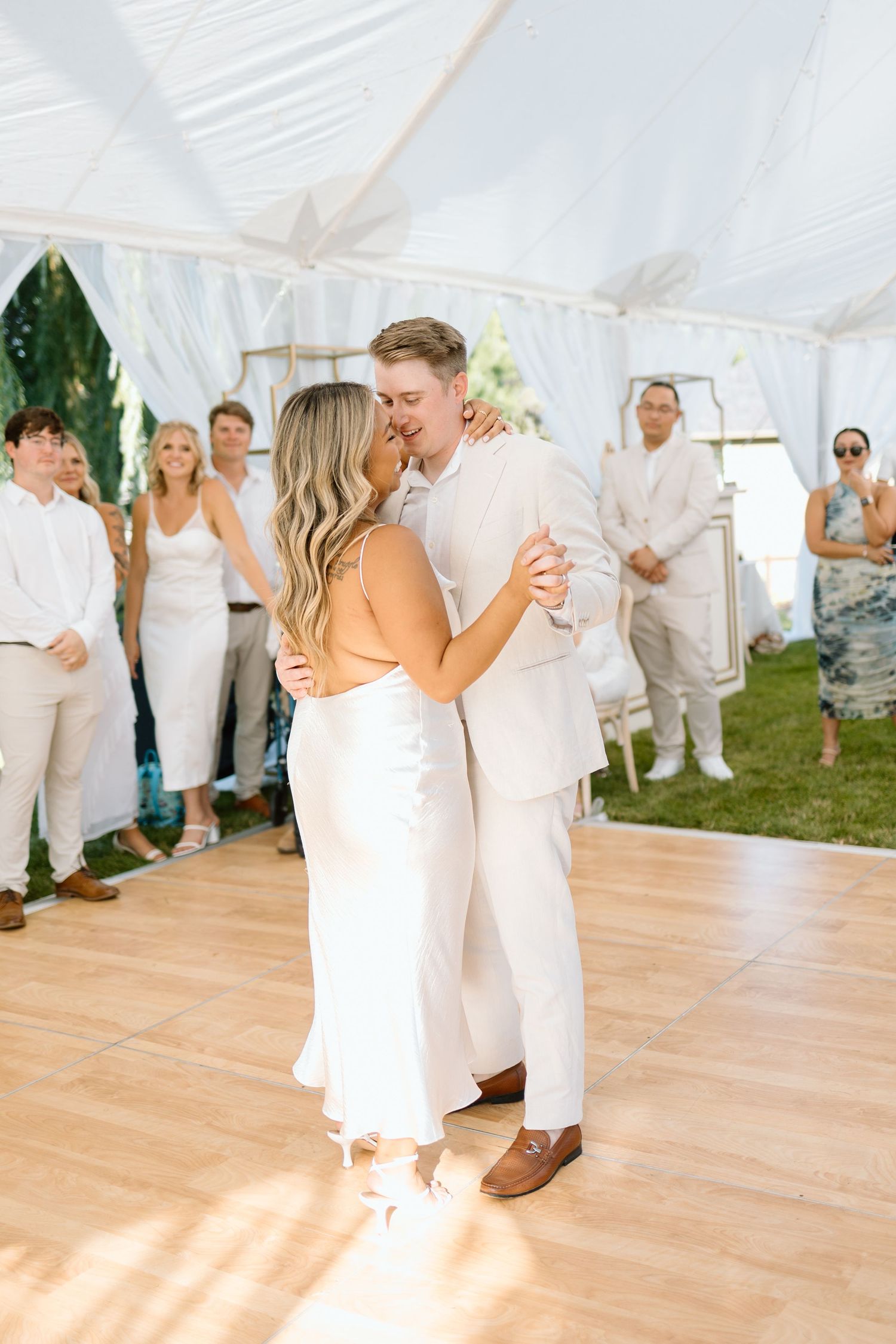 People in white attire gather at an elegant outdoor tent wedding reception as a couple shares a dance on a wooden floor.