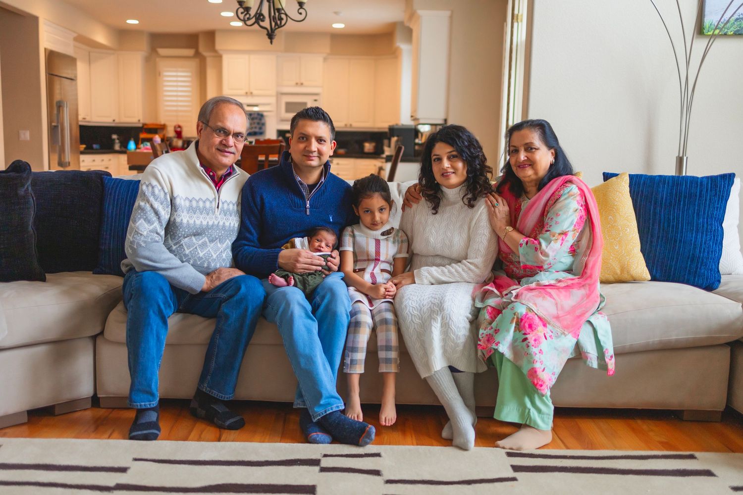 Multiple generations sit together on a beige couch in a modern living room with blue accent pillows.