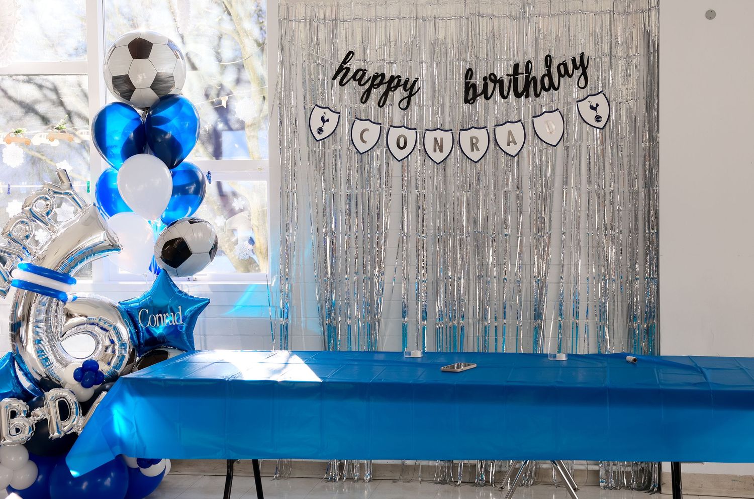 Blue and silver birthday party decorations with balloons football ball shaped and a name and happy birthday banner against a white backdrop.