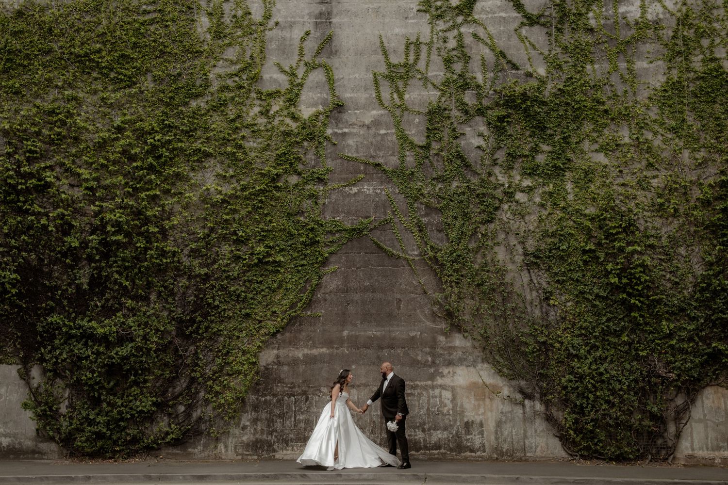 A bride and groom hold hands and laugh in front of a stone wall covered in ivy.