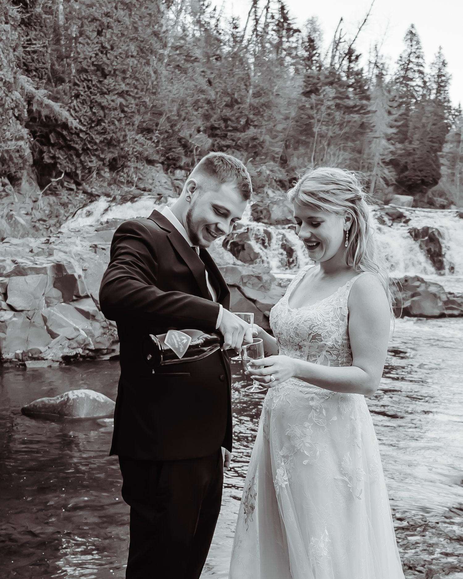 A black and white wedding photo captured by a waterfall with evergreen trees in the background.