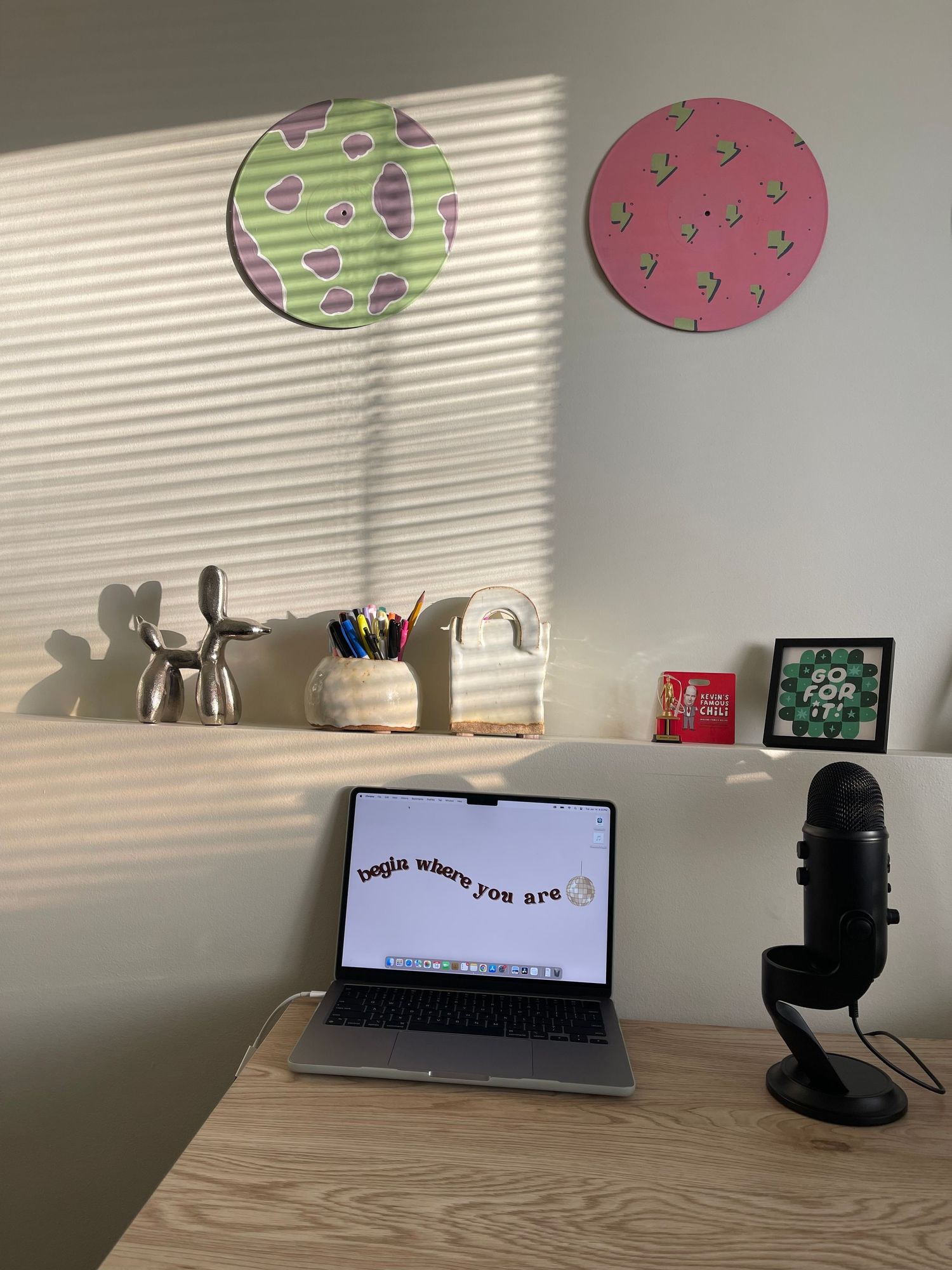 A minimalist desk setup with a laptop, microphone, and decorative circular wall art in green and pink casting window shadows.