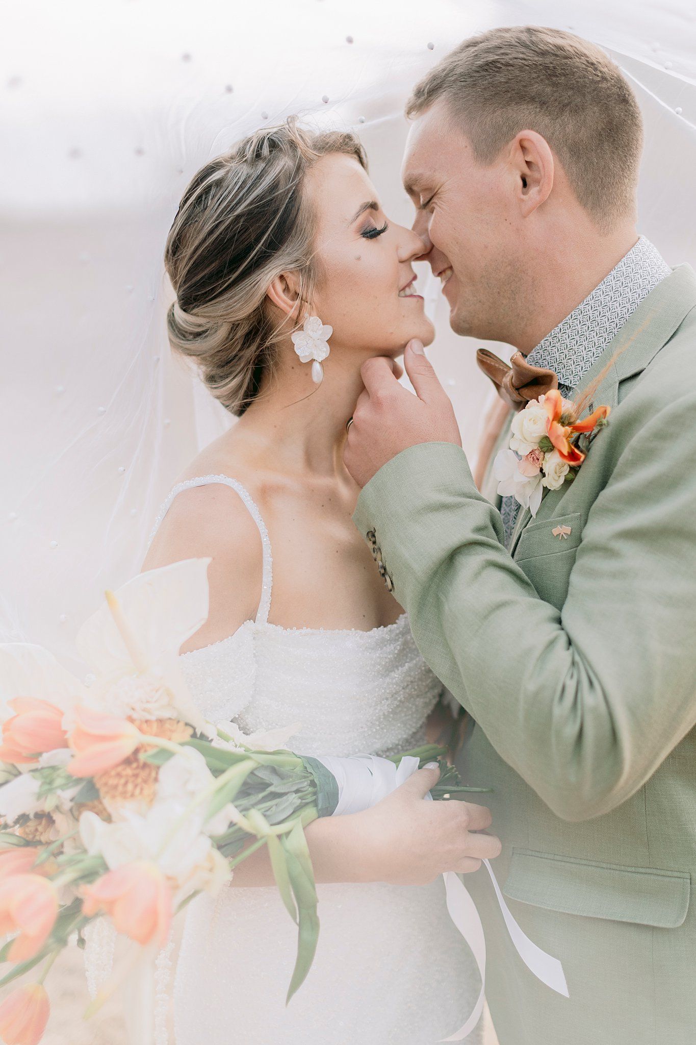 Bride and groom sharing an intimate moment under a veil, groom in sage green suit with floral boutonniere.