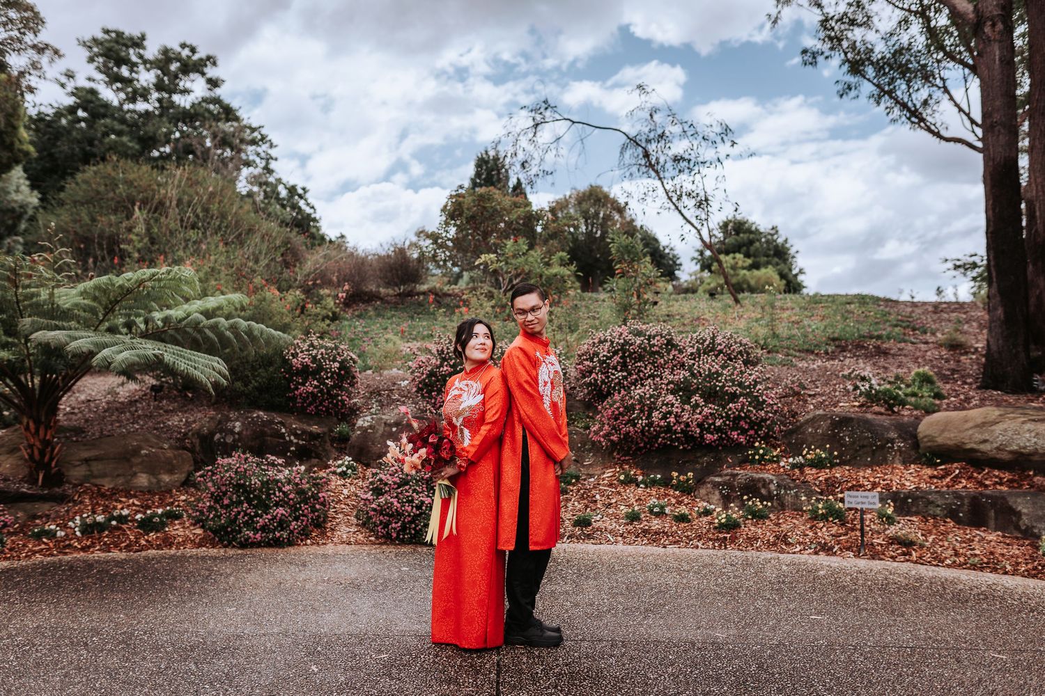 Two people in vibrant red traditional attire stand together in a lush garden setting with ferns and flowering shrubs.