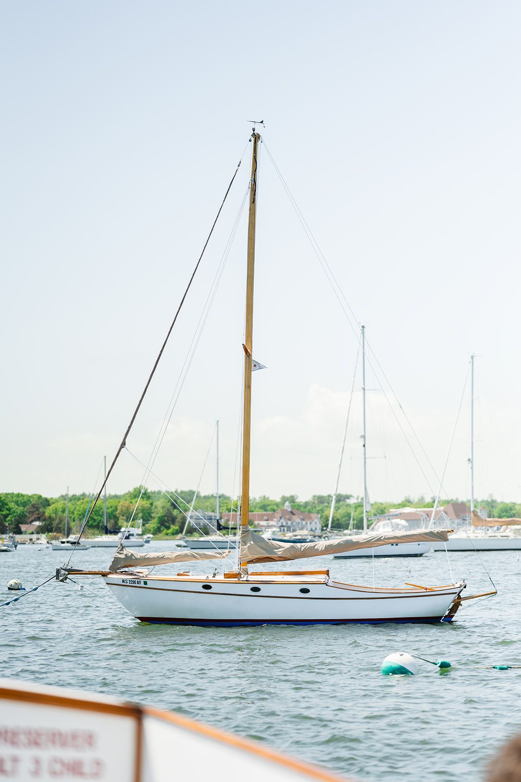 A classic sailboat with wooden mast anchored in a peaceful harbor with other boats visible in the background.