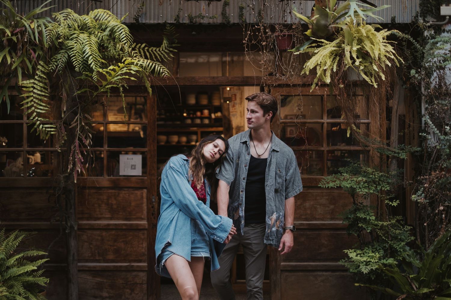 Stylish couple holding hands outside a rustic shop surrounded by lush tropical plants.