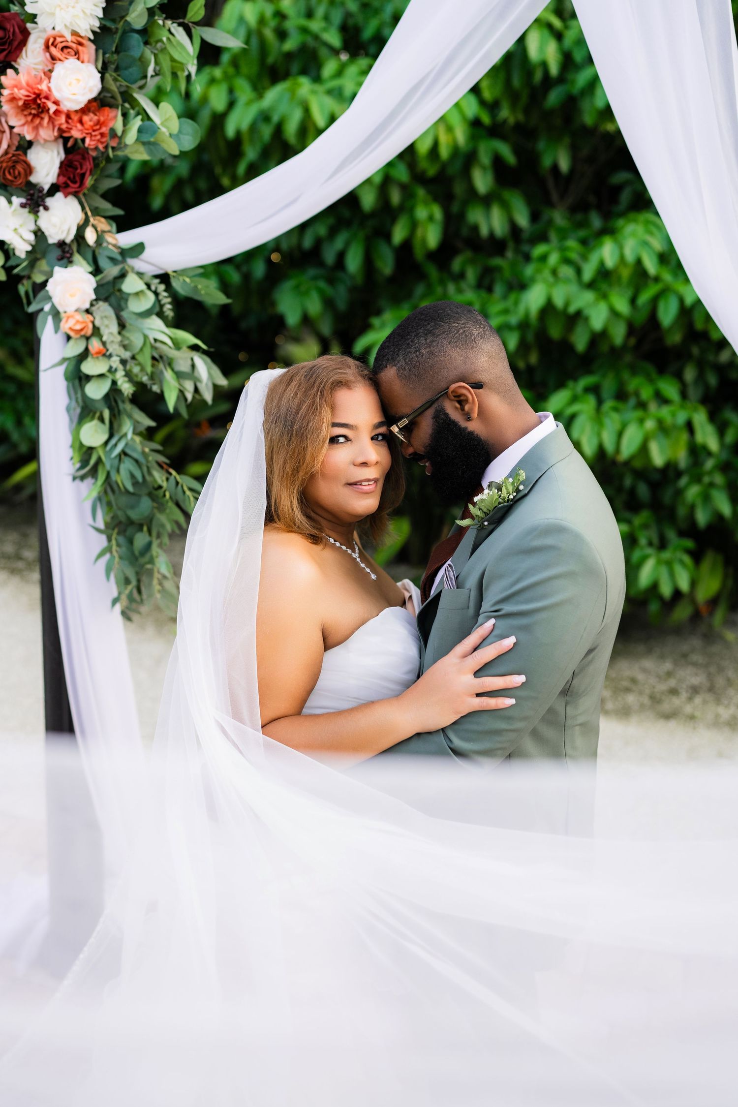 A romantic moment captured under a white draped wedding arch decorated with coral flowers and greenery.