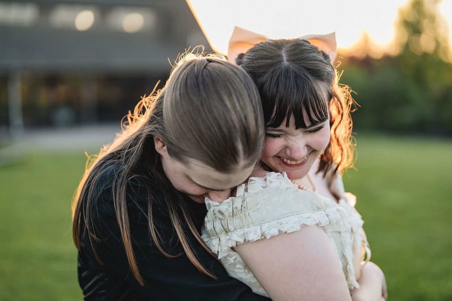 Two women share a joyful moment together outdoors under willow trees by a lake, one wearing a white lace dress.