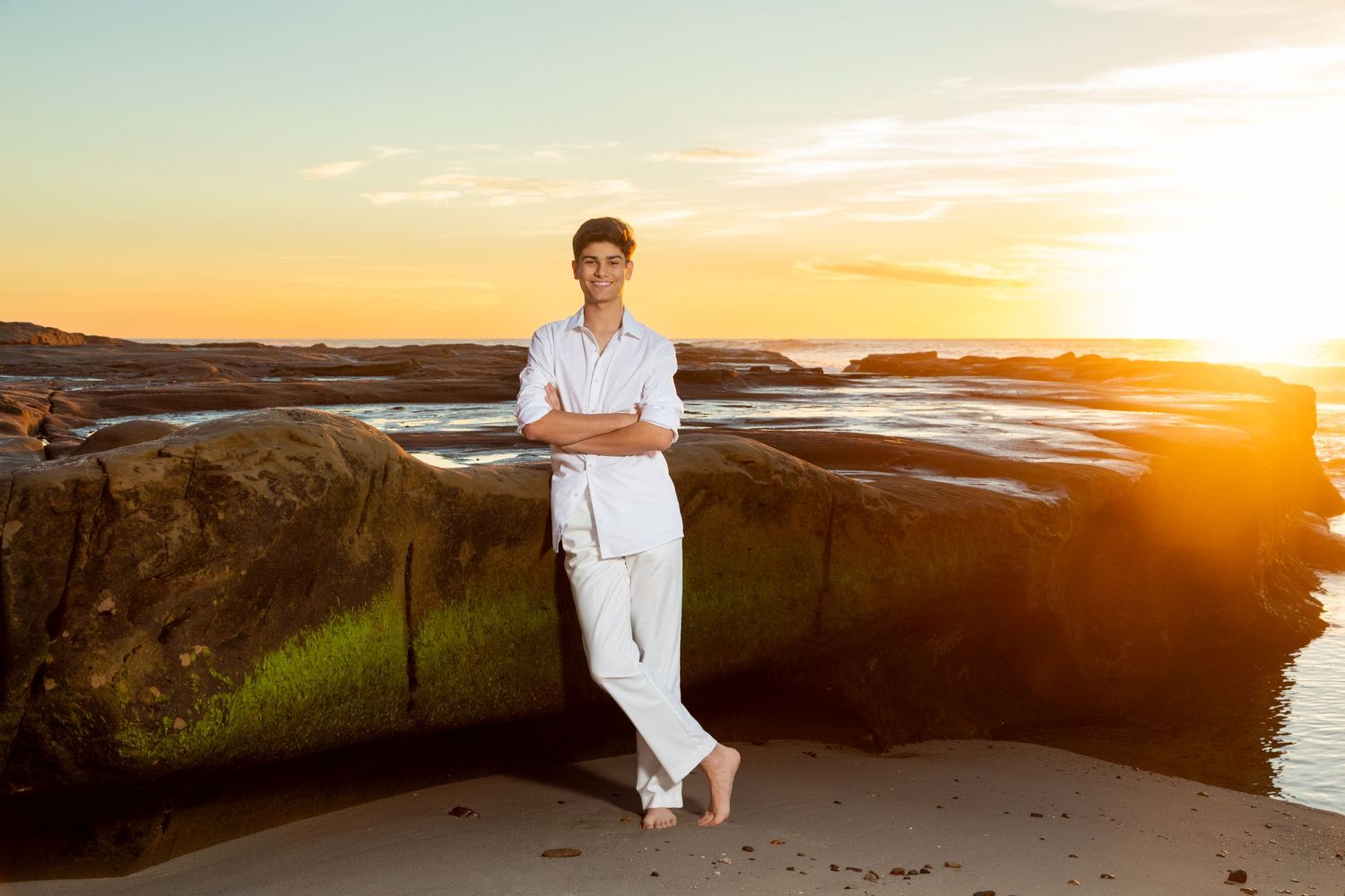 Figure in white clothing leans against rocky beach formation during sunset.