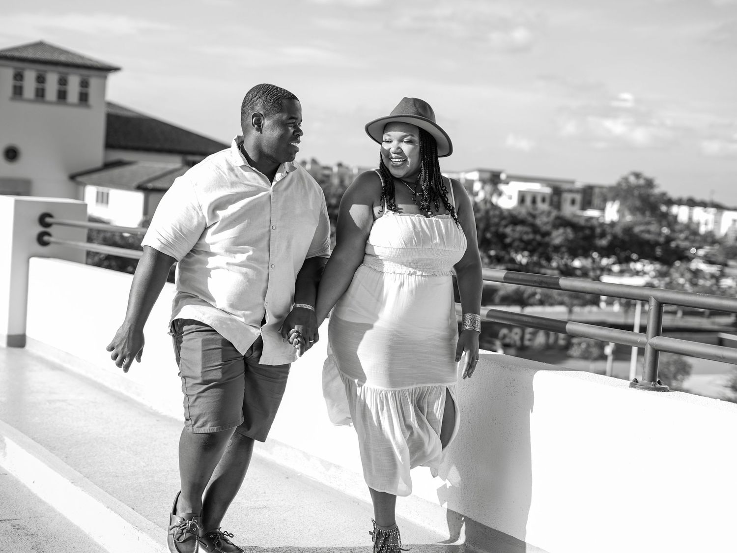 A couple walks along a waterfront promenade with buildings in the background.