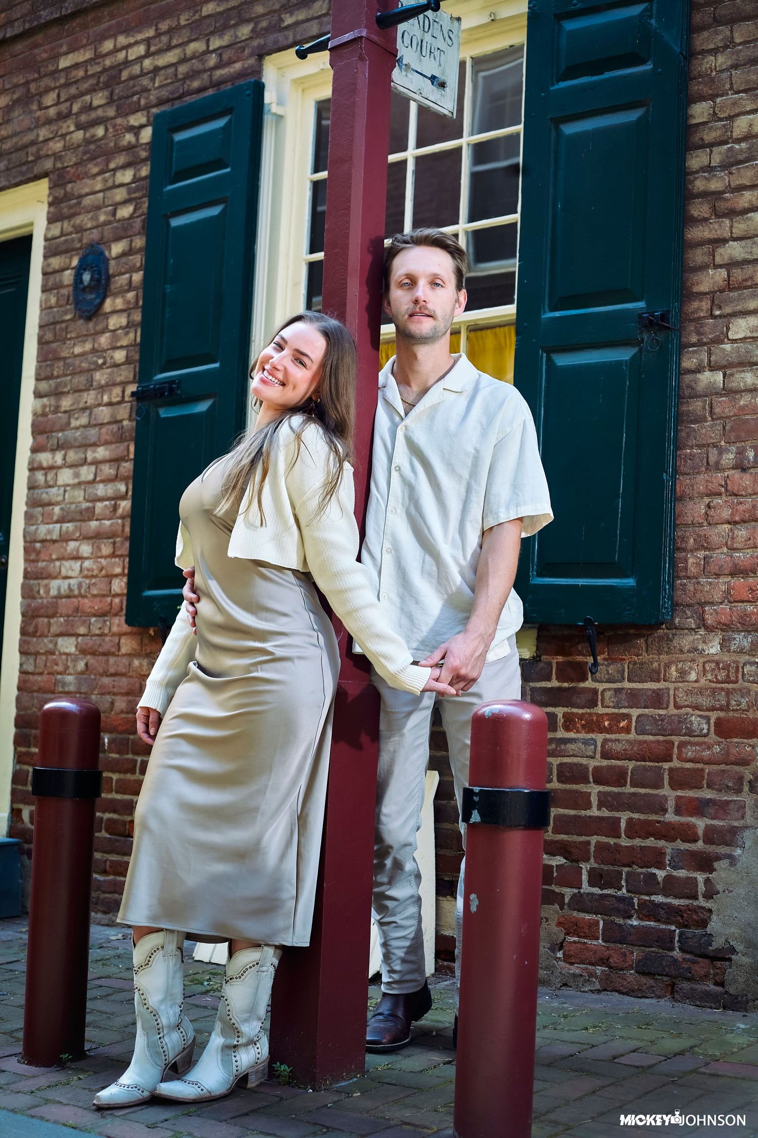 A romantic couple in white outfits poses against a brick wall with green shutters.