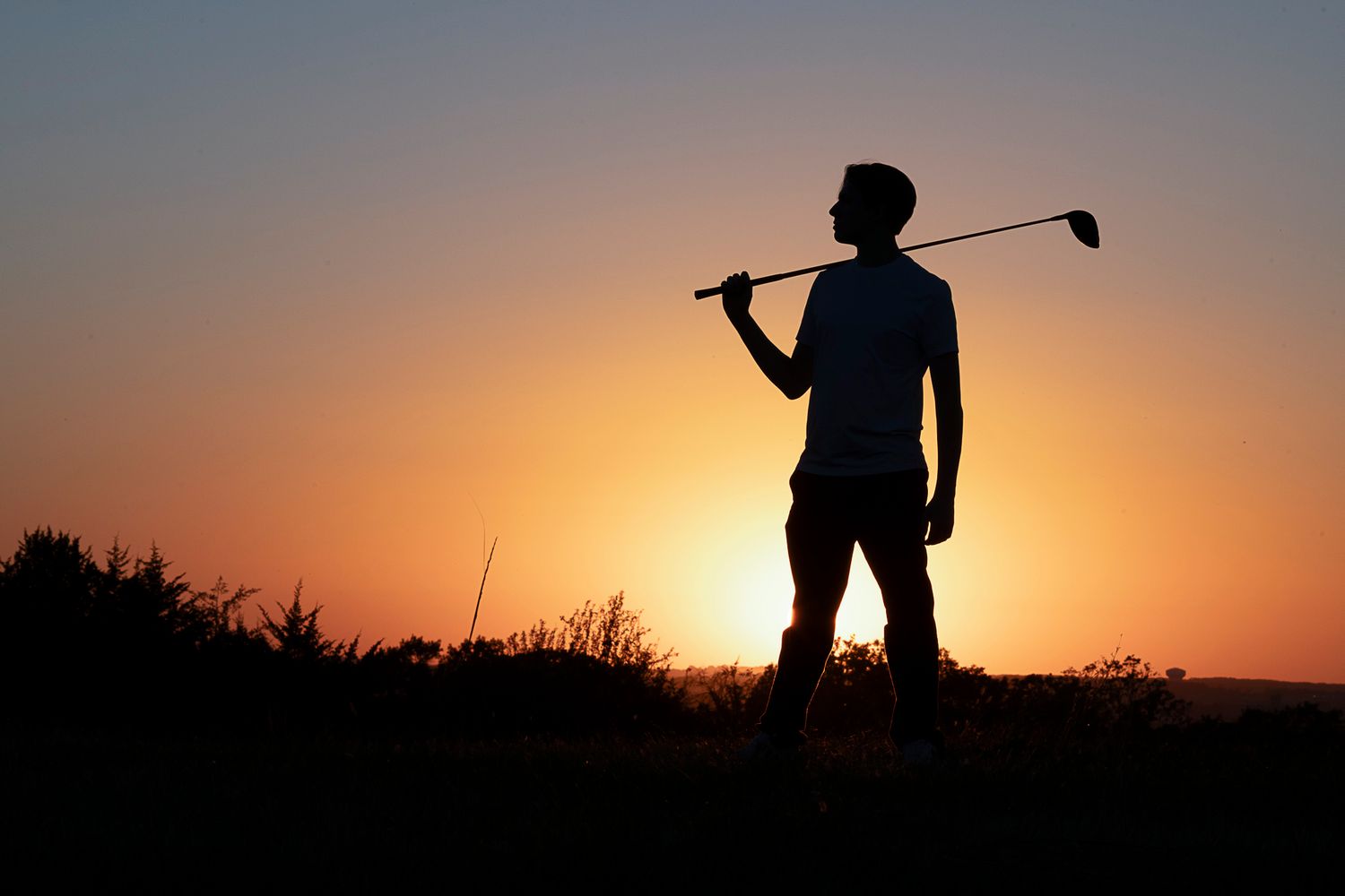 A silhouetted golfer stands against a vibrant orange sunset sky while holding a golf club.