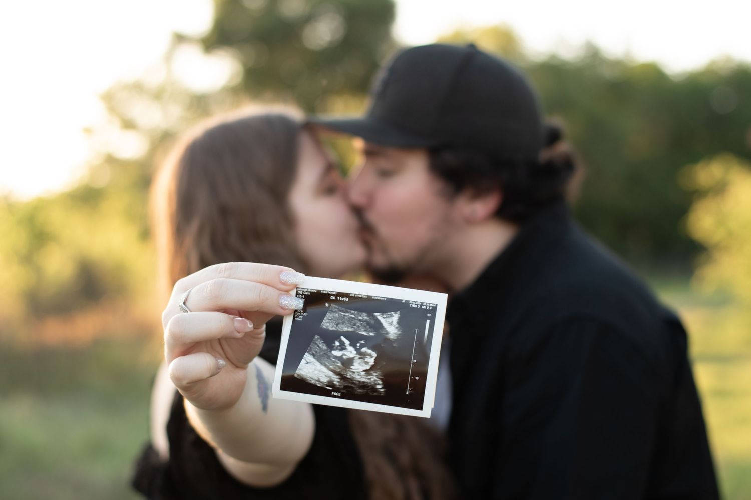 Couple holding ultrasound photo while sharing a kiss in sunset light.