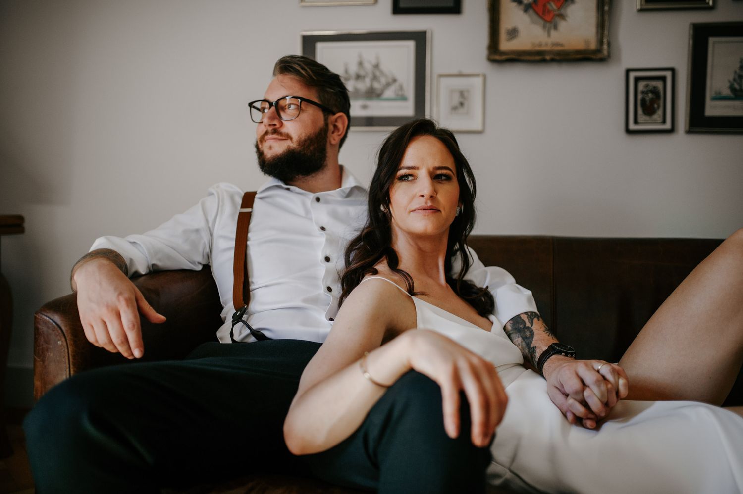 Stylish tattooed bride and groom relaxing on leather sofa in bohemian indoor setting.