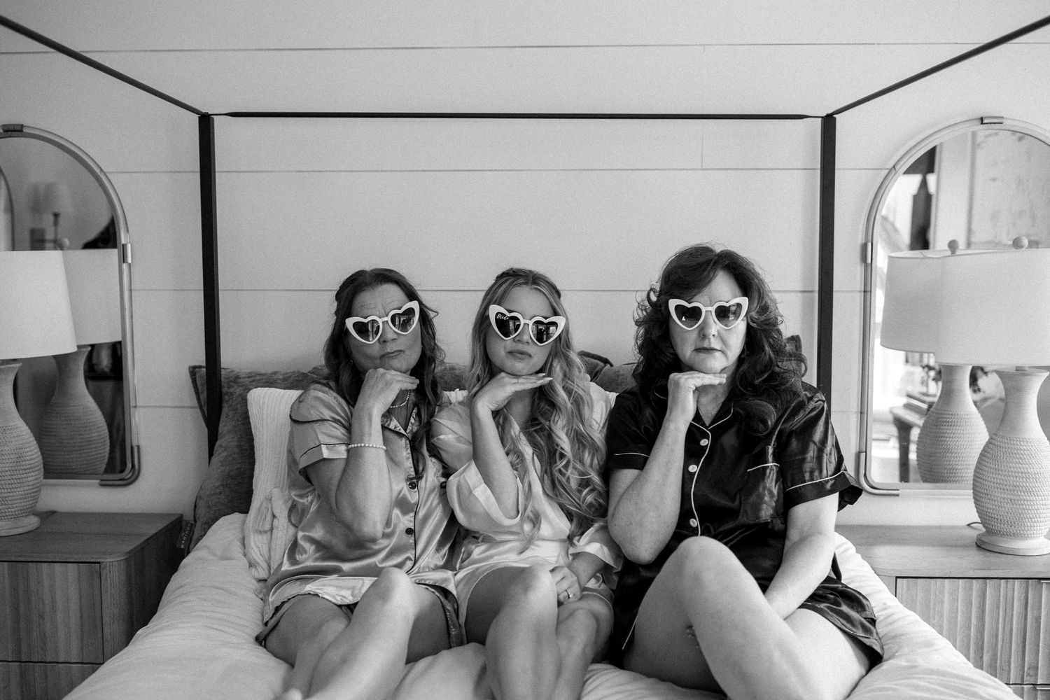 Three friends wearing sunglasses pose together on a white bed.