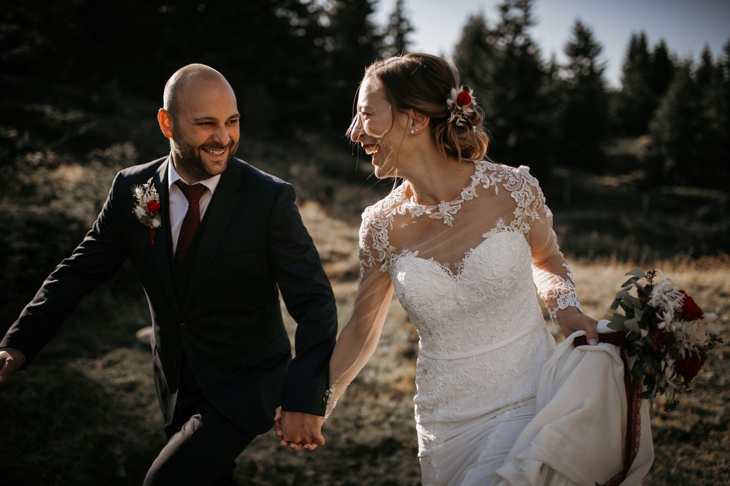 Laughing bride and groom running hand in hand through golden autumn mountain meadow.