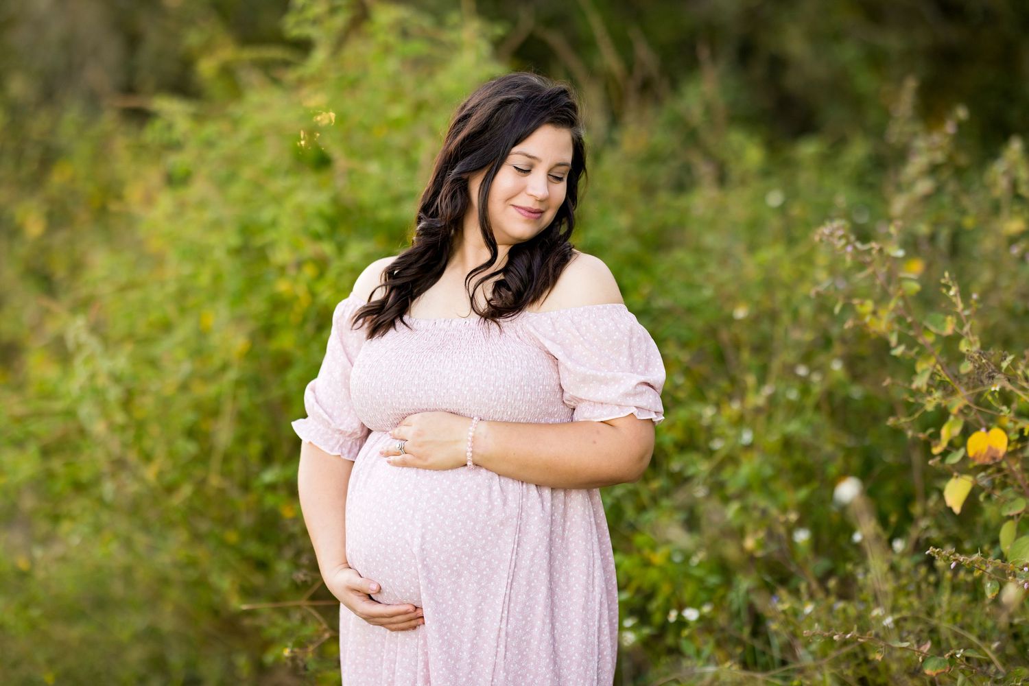 Pregnant woman in pink dress poses in lush garden setting during maternity photoshoot.