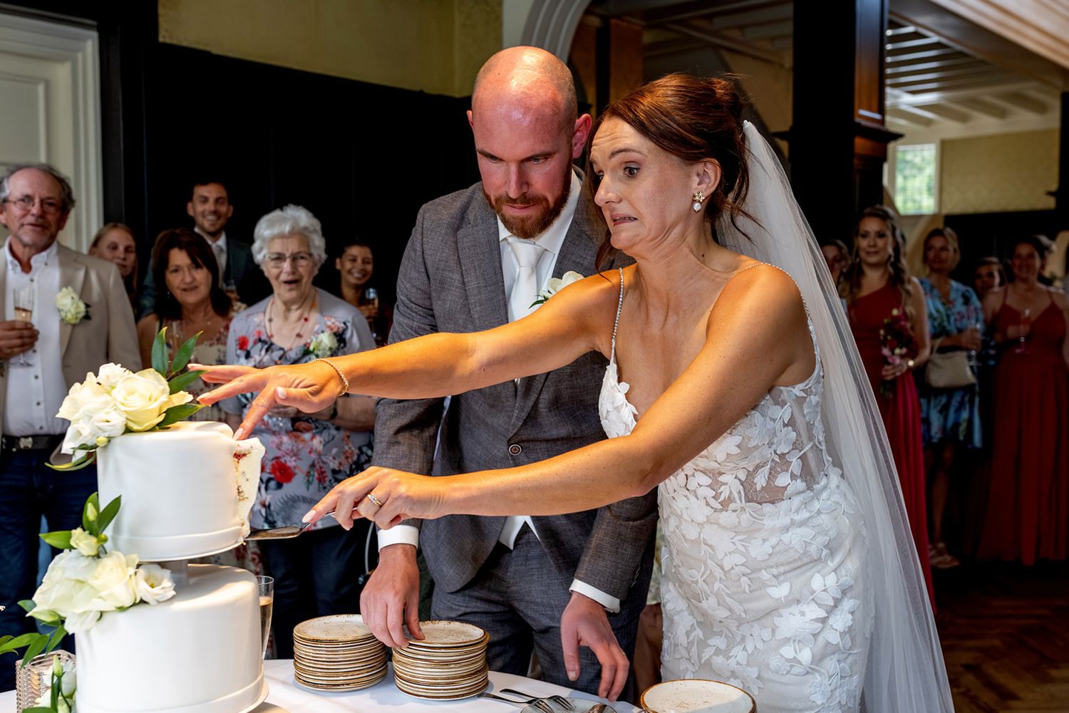 De bruid snijdt de witte taart, gedecoreerd met witte rozen aan. Ze probeert de taartpunt op het schoteltje te krijgen. Ze kijkt gespannen