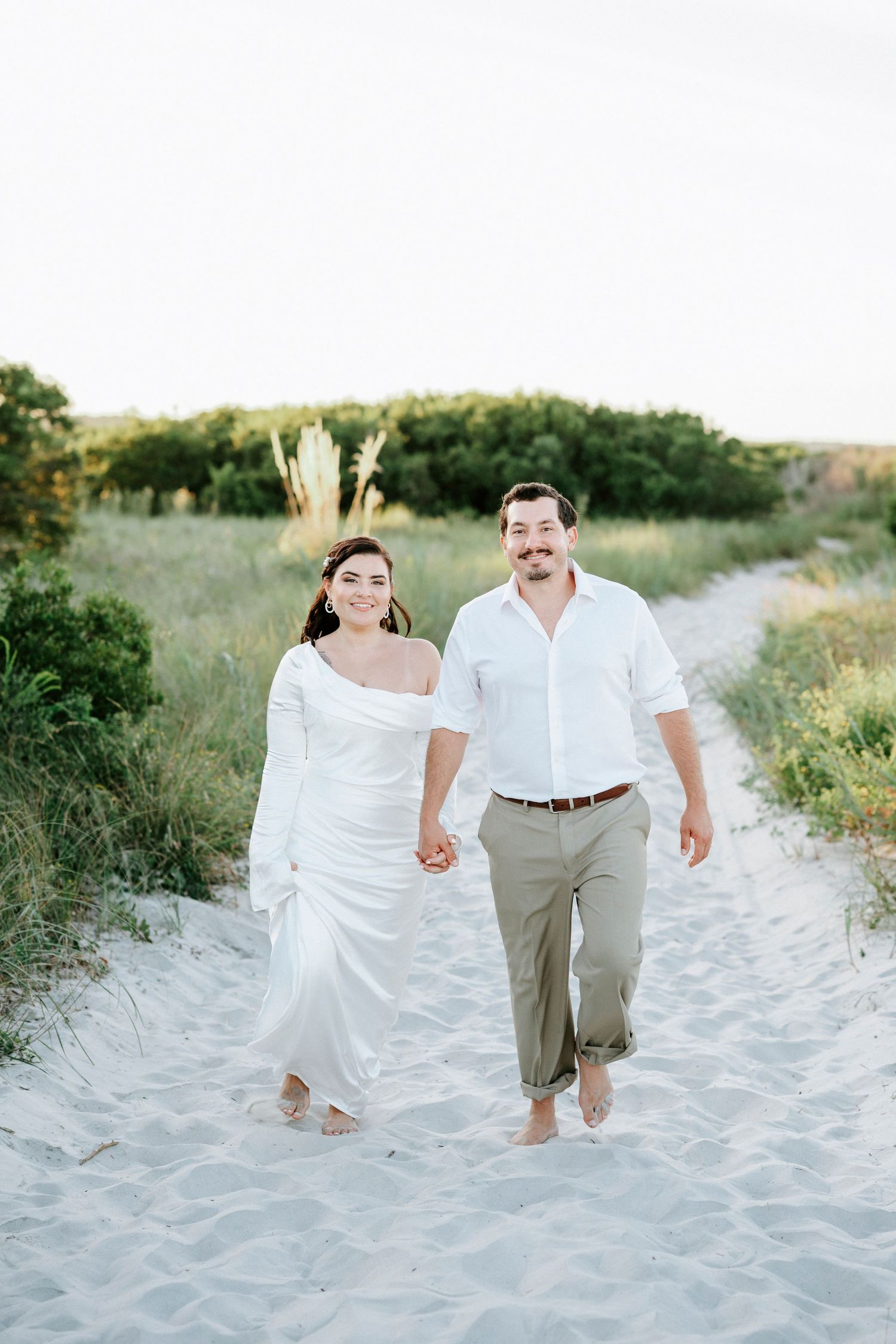A couple in white clothing walks together along a sandy beach path during a sunny afternoon.