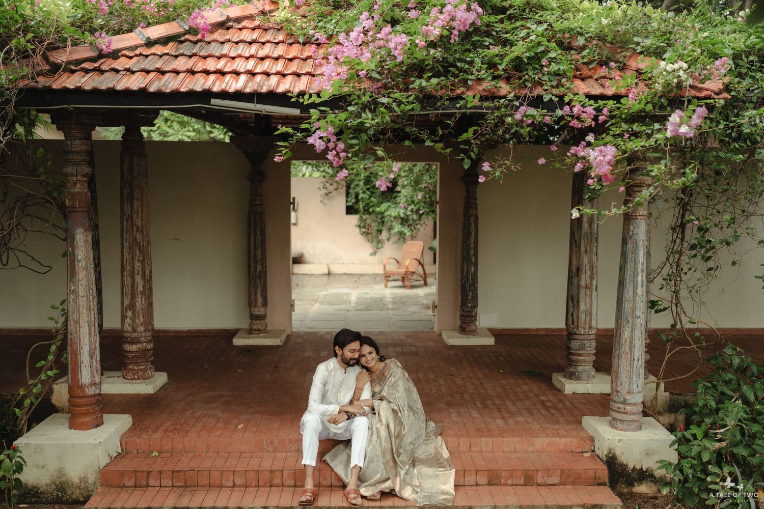 A covered walkway with red tile roof and flowering vines leads to a traditional Mediterranean style building.