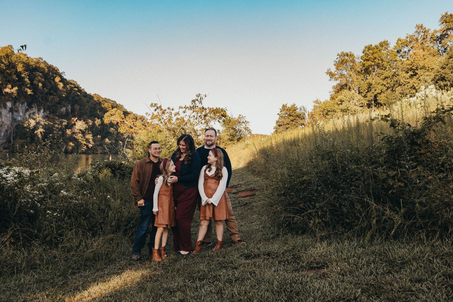 Family of five posing together on a grassy hillside path surrounded by autumn foliage and a rocky bluff.