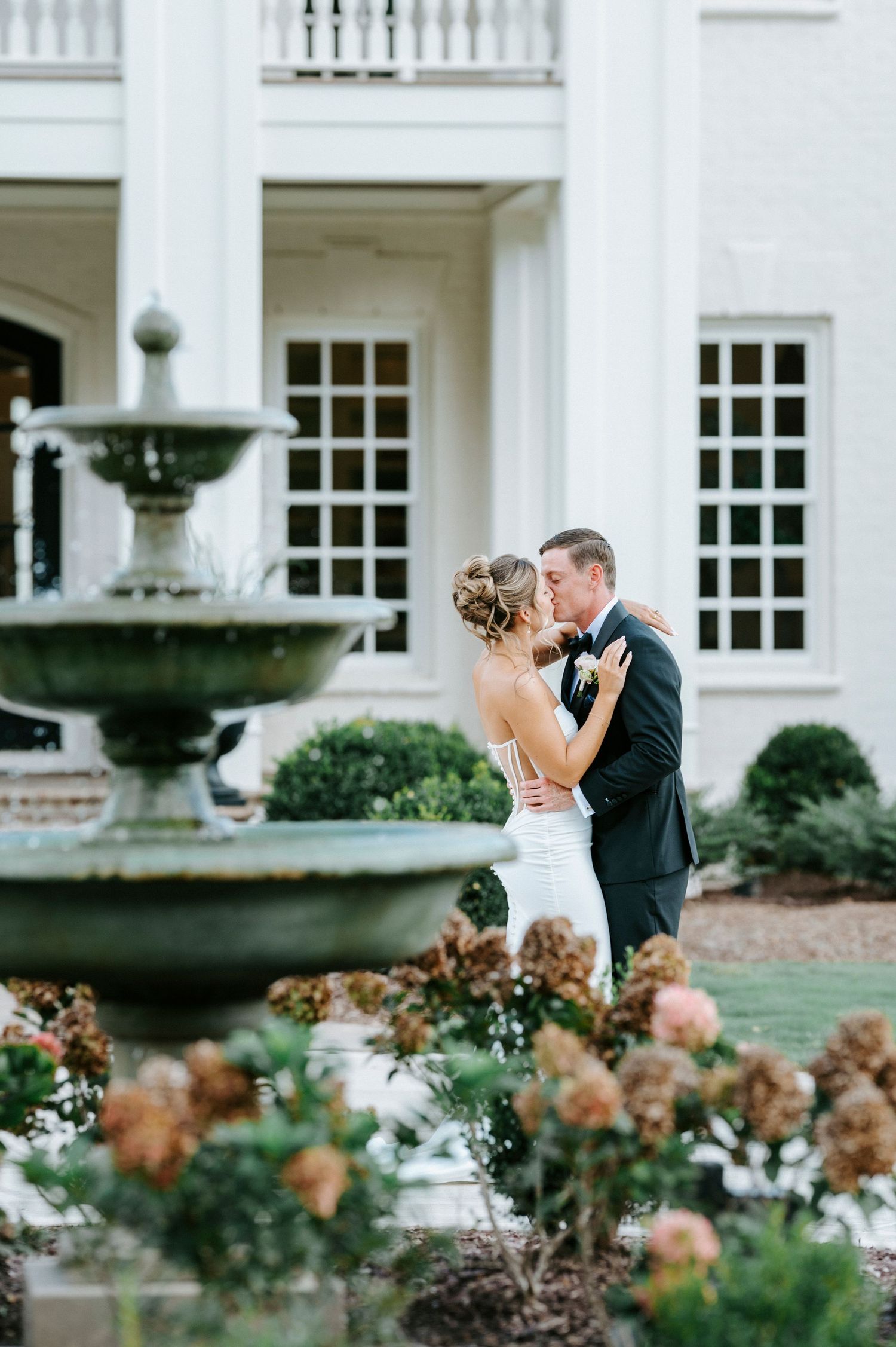 Romantic kiss between newlyweds in formal attire by an elegant white mansion with fountain and floral landscaping.