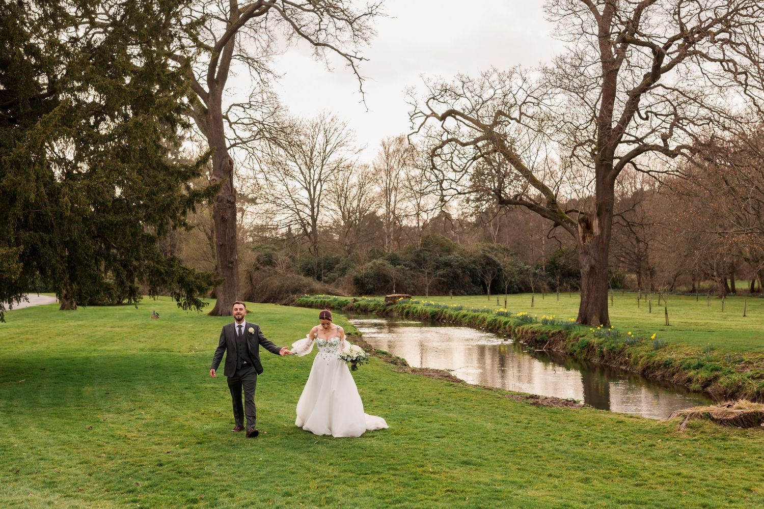 A couple walks through a scenic park with trees and a winding stream on their wedding day.