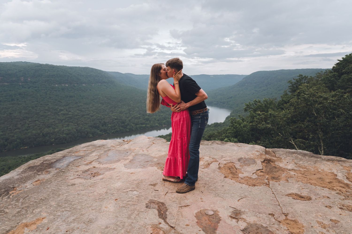 Two people share an intimate moment on a rocky cliff overlooking a scenic mountain valley and winding river.
