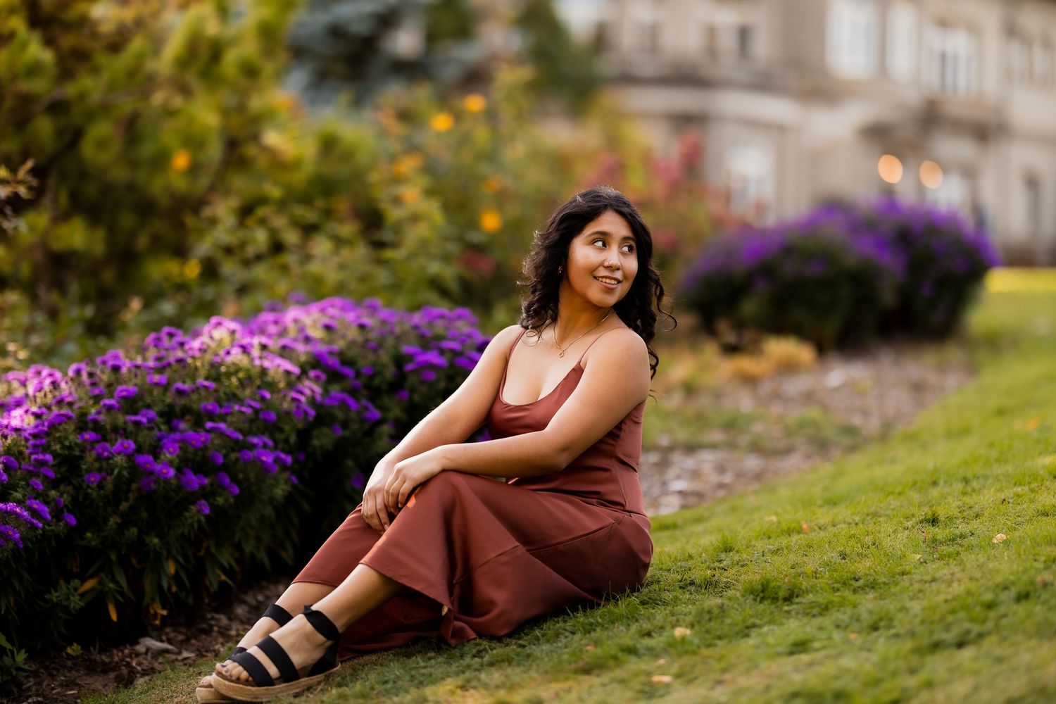 A person in a brown dress poses on a grassy slope with vibrant purple flowers in the background.