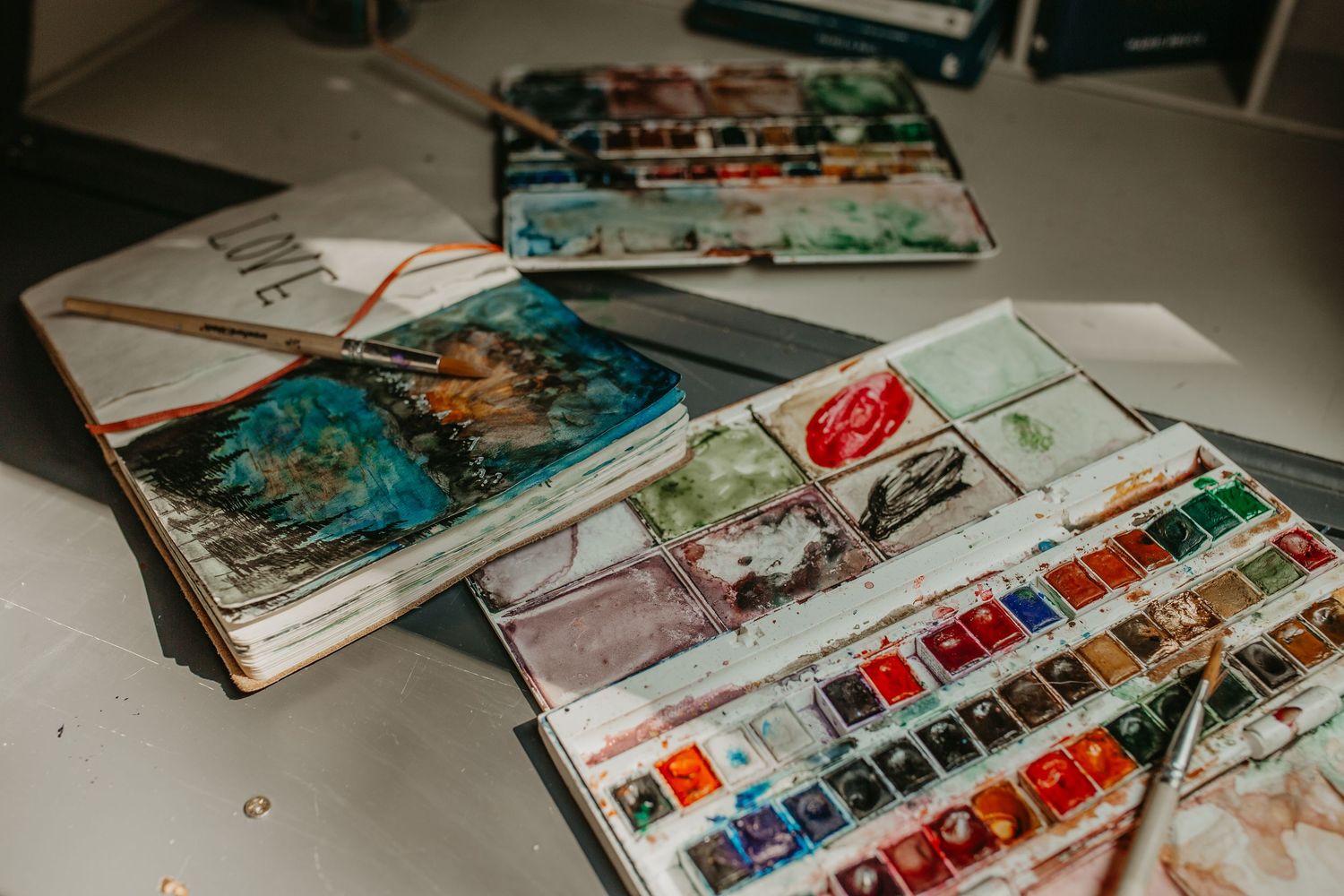 Open watercolor paint sets and sketchbooks scattered on a white surface with vibrant paint palettes visible.