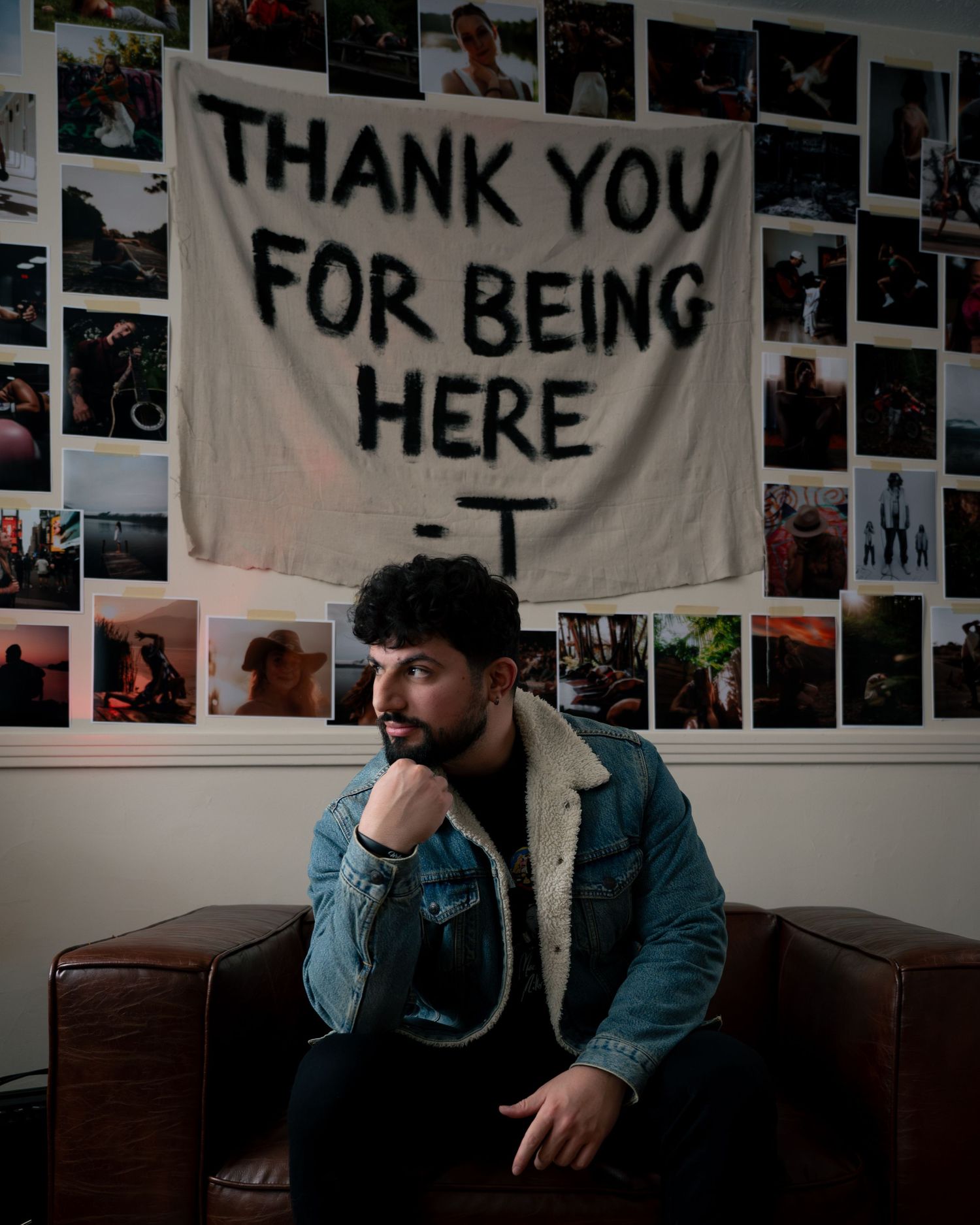 A contemplative figure sits on a leather couch beneath a white banner reading 'THANK YOU FOR BEING HERE' with photos on the wall.