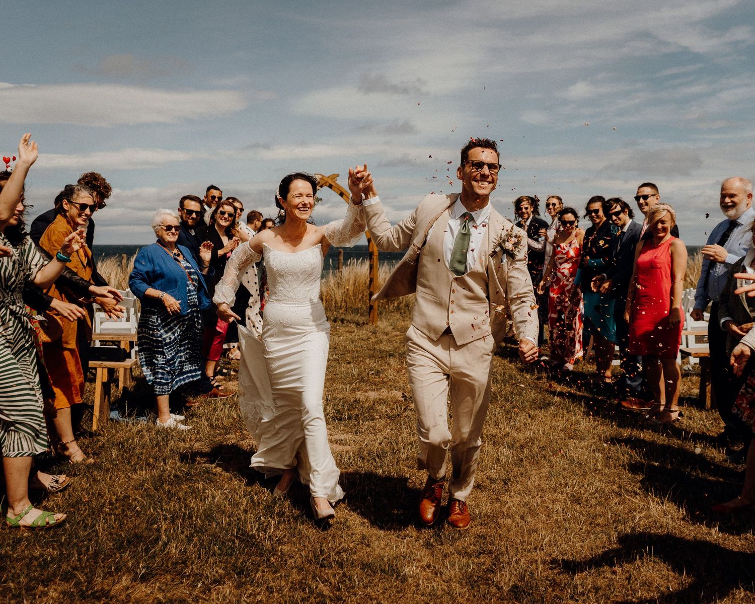 Wedding guests line up outdoors on a grassy hillside overlooking the ocean during a sunny summer celebration.