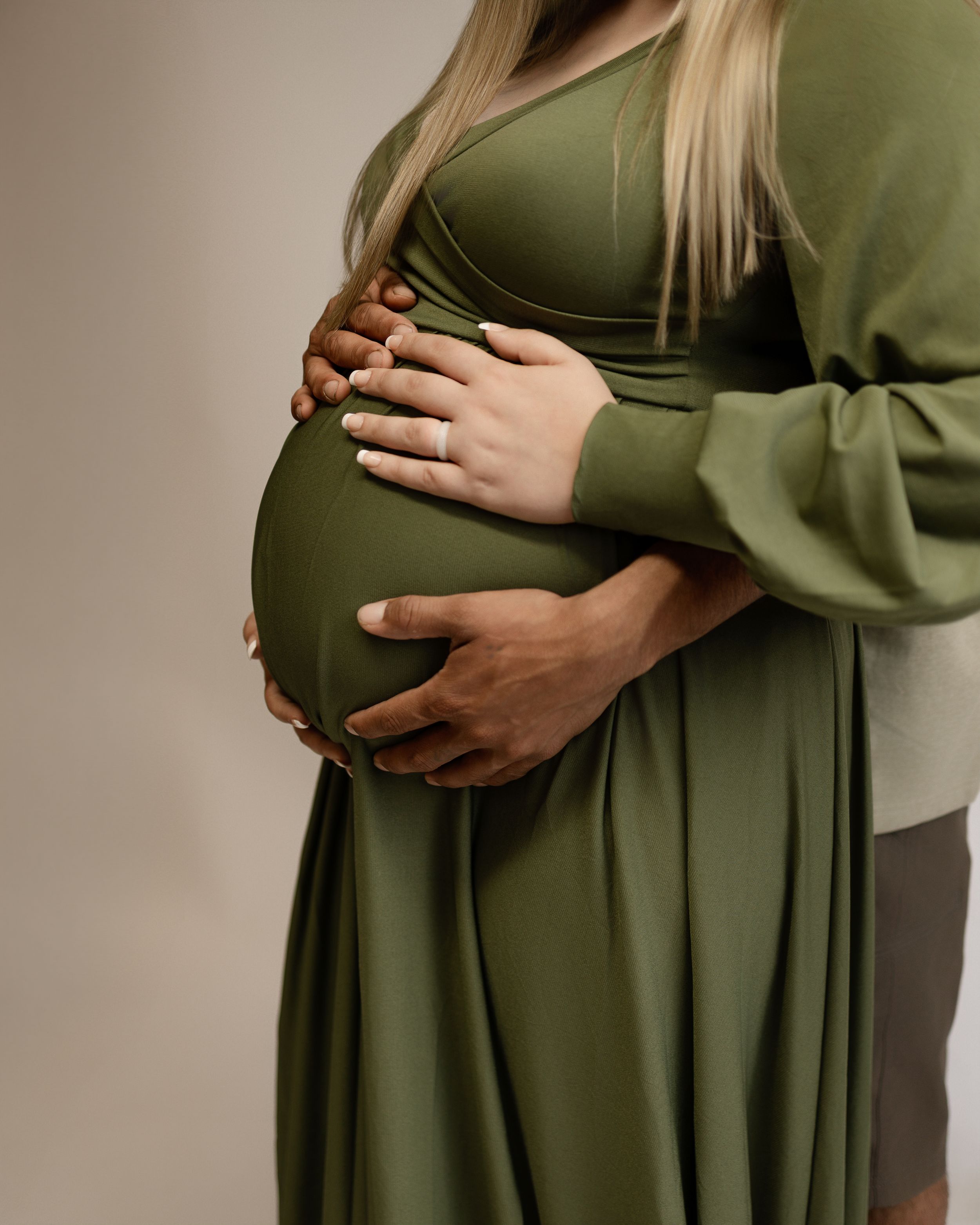 Hands gently cradle a pregnant belly wearing an olive green maternity dress against a neutral background.